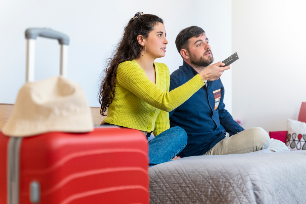 hotel guests sitting on guest room bed watching TV, with a suitcase in the foreground